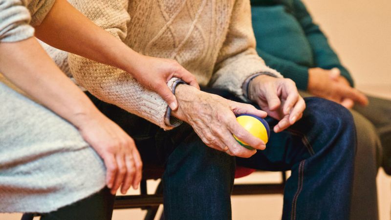 Una persona mayor sostiene una pelota de colores mientras su cuidadora le coge del brazo